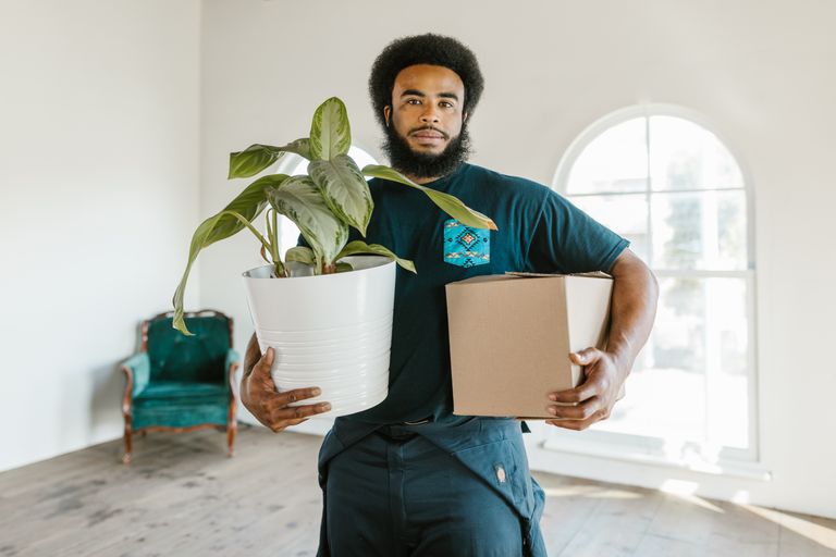 A man in a crew neck t-shirt holds a potted plant and a box in a bright, indoor setting.